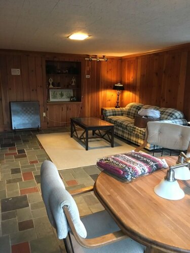 Cozy living room with wood-paneled walls, comfortable plaid sofa, armchair, and a central coffee table ideal for relaxation and gatherings.