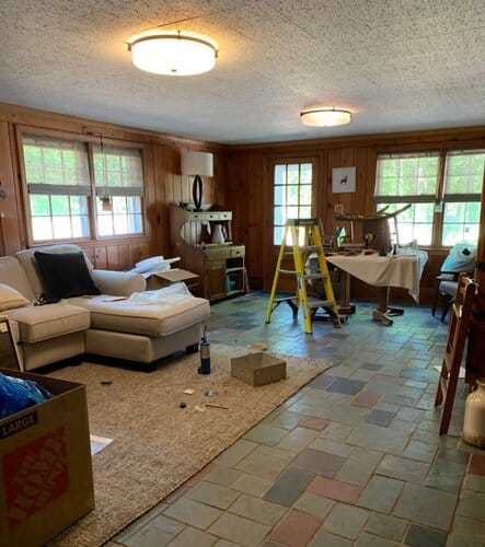 1.Cozy living room with wooden panel walls, large windows, and a mix of furniture including a sectional sofa, dining table, and craft area with supplies and a ladder.