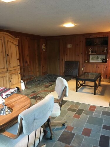 Bright cozy living room with wooden walls and tiled flooring, featuring comfy chairs, a coffee table, and decorative shelves, perfect for family gatherings or relaxation.