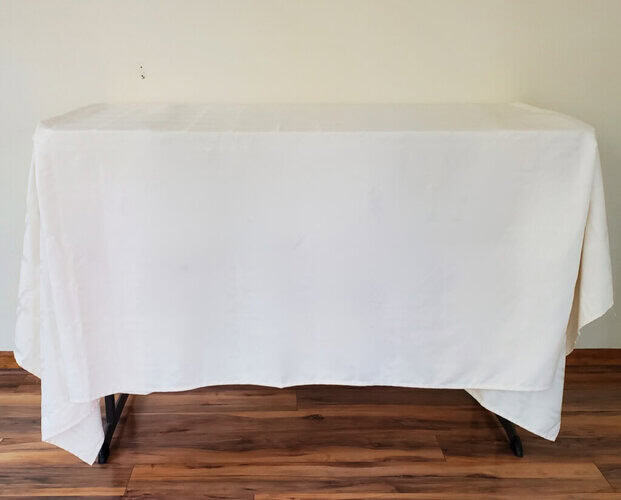 White craft table with black metal legs covered in a white cloth, set against a beige wall and wooden floor, perfect for arts and crafts or creative projects.